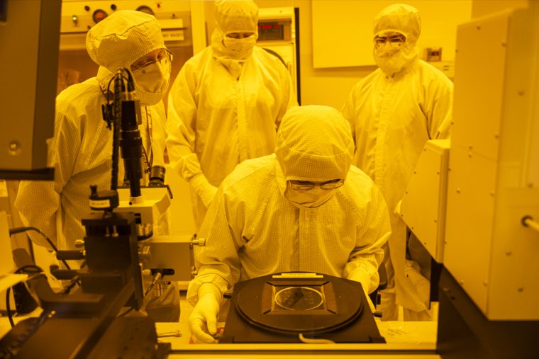 Four people in cleanroom suits gathered around a tool, watching a wafer being loaded