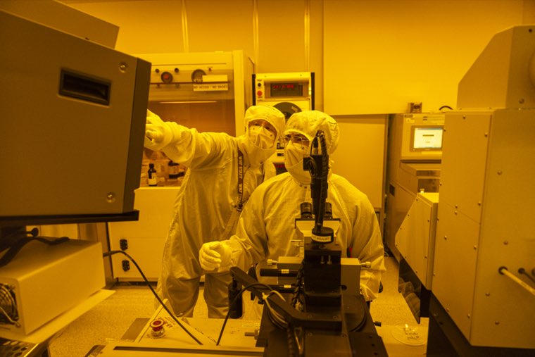 Two researchers working at a tool in the yellow-lit lithography bay